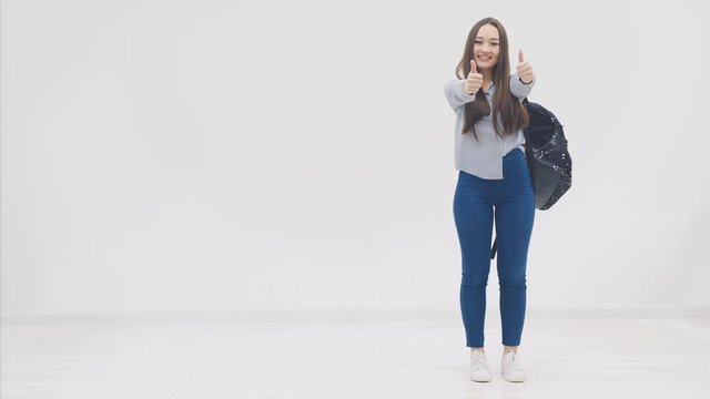Happy, smiling, full height confident girl with long hair appearing on a white background with backpack behind her shoulders, looking to the side, giving thumbs up.