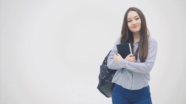 An asian schoolgirl appearing on a white background with a backpack slung over her shoulder, holding book near chest, posing.