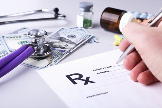 Male doctor or pharmacist holding jar or bottle of pills in hand on a background of dollars banknotes and writing prescription on a special form. medical costs and healthcare payment concept.