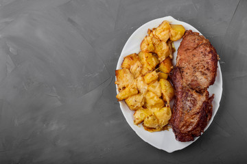 Pork loin steak and potatoes baked with cheese on a white plate, gray background