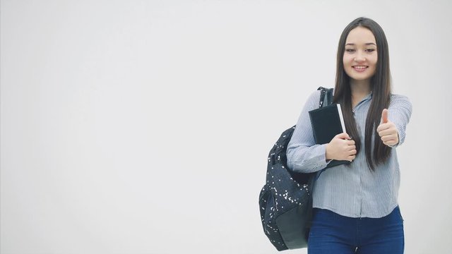 An asian schoolgirl standing with a backpack slung over her shoulder, holding book near chest, giving thumbs up.