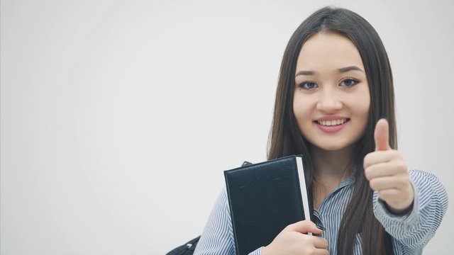 An asian schoolgirl standing with a backpack slung over her shoulder, holding book near chest, giving thumbs up.