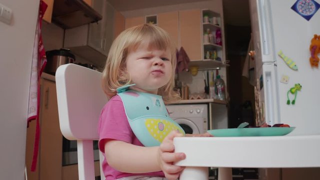 Little two years old blond blue-eyed girl grimaces while having her dining, 4K shot