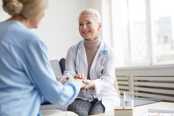 Fototapeta premium Portrait of mature female doctor holding hands on senior woman and smiling happily during consultation in medical clinic, copy space