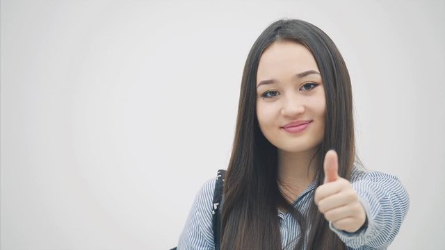 An asian schoolgirl standing with a backpack slung over her shoulder, holding book near chest, looking to the side, giving thumbs up.