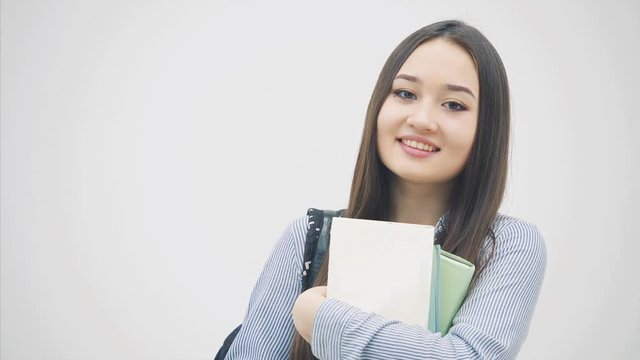 An asian schoolgirl appearing on a white background with a backpack slung over her shoulder, holding a stack of books near chest, posing, smiling.