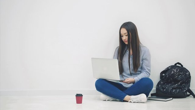 Lovely young asian lady sitting on the floor in lotos position, searching in the internet, waving hello. School supplies around her.