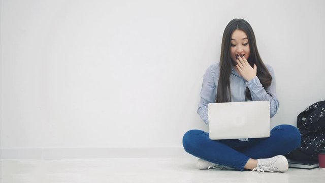 Lovely young asian lady sitting on the floor in lotos position, searching in the internet, covering her mouth by a hand surprised with something.