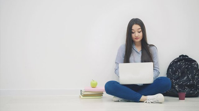 Asian lady sitting on the floor in lotos position, with laptop, taking pen and notebook out of her bag, writing something down.