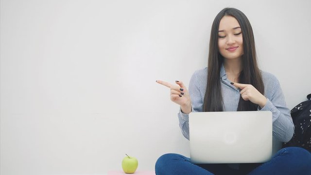 Lovely young asian lady sitting on the floor with laptop, in lotos position,pointing her fingers to the side.