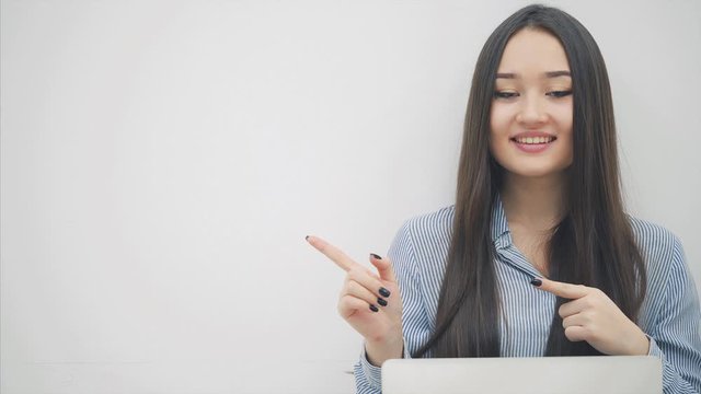Lovely young asian lady sitting on the floor with laptop, pointing her fingers at the camera, then to the side.