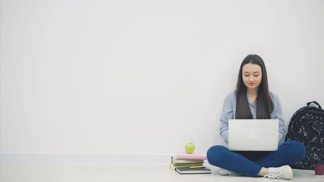 Lovely young asian lady sitting on the floor in lotos position, searching in the internet, waving hello. School supplies around her.
