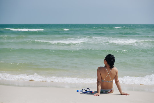 Young woman in bikini sitting on beach and looking at sea waves after snorkeling
