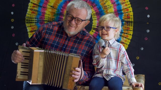 blond child and father with gray hair and beard in plaid shirts celebrate together family holiday. Father plays the accordion while boy holds a microphone. Lovely family duet