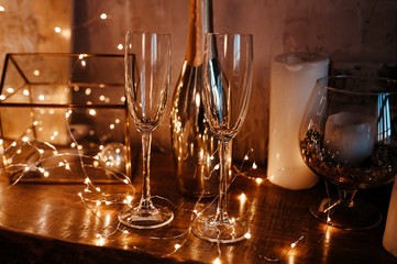 two empty glasses and a golden bottle on a wooden table in garlands and bulbs in the dark, table setting for a romantic evening for two lovers