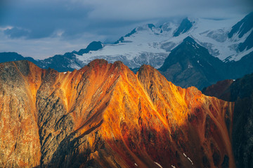 Atmospheric alpine landscape with red rockies in golden hour. Scenic view to big orange rocks and giant snowy mountains with glacier in sunrise. Wonderful highland scenery. Flying over mountains.