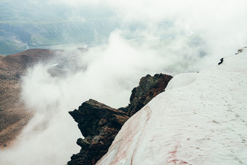 Fototapeta premium Beautiful pointy cliff on high rocky mountain edge with snow among thick low clouds. Atmospheric minimalist alpine landscape. Pointed craggy stone near abyss in clouds. Wonderful highland scenery.