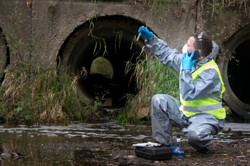 scientist in protective suit, gloves and mask, holding a vial of liquid and is calling on mobile phone user to report results of samples from the river, amid the purifying pipe