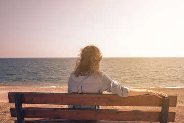 Rear view of young woman wearing blue casual dress sitting on a bench while looking away to horizon and sunset over sea