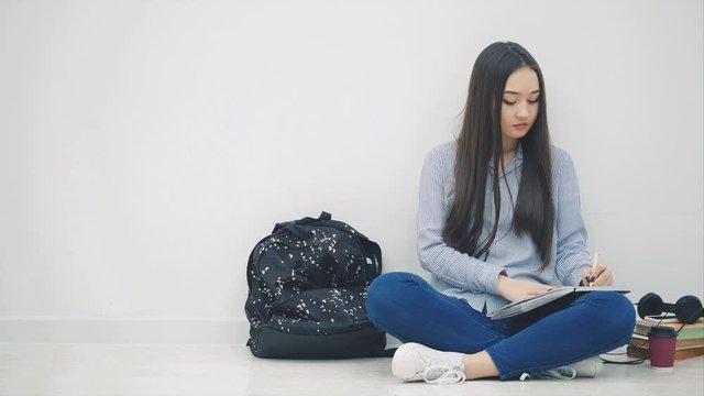 Inspired young asian girl sitting on the floor, finished writing, takes phone and headphones and is listening to music, dancing.