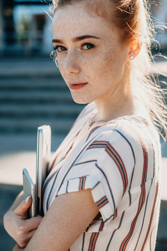Close up portrait of a charming woman with red hair and freckles looking seriously at camera against sunset holding a laptop and a smartphone.