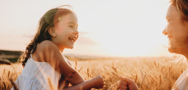 Side view portrait of a happy little girl laughing while looking at her mother in a field of wheat against sunset.