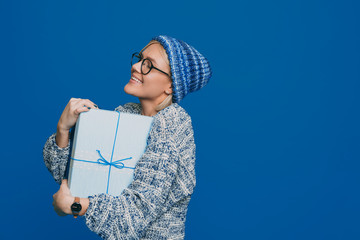 Portrait of a charming young blonde woman embracing a blue gift box laughing against a blue background. Birthday or new year celebration concept.