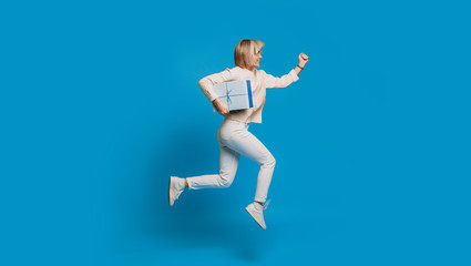 Fototapeta na wymiar Side view portrait of a beautiful young woman running with a blue gift box against a blue background. Fast delivery concept.