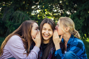 Three young attractive woman sharing secrets sitting on green grass in the park. Cheerful...