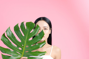 Asian woman holding a tropical leaf with natural skin on pink background. Beauty concept.