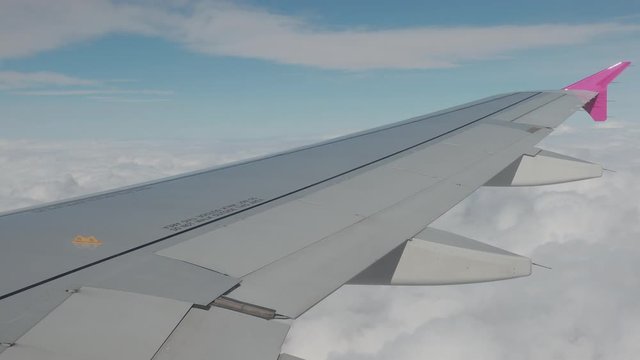 Amazing view on plane wing of grey, pink colours. Behind airplane there are lots of white fluffy clouds, bright day blue sky.