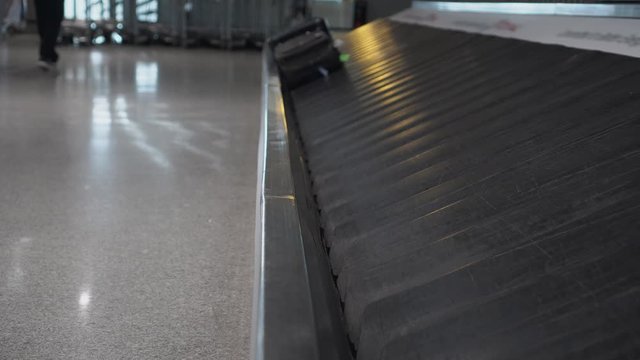 Two suitcases, brown and black, are approaching on a conveyor belt at the airport. On one of the suitcases, a green tag is attached. Silhouettes of passing people in the background. Travel concept. 4K