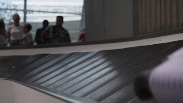 Purple suitcase and a black bag are moving along the conveyor belt at the airport. Close-up. Rounding, bending the conveyor belt. Silhouettes of people waiting for their luggage in the background. 4K