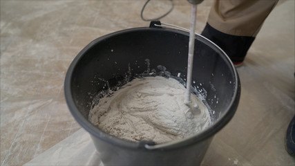 walls of the apartment using an electric drill, close-up. Worker mixes construction putty in a bucket. The white building mix is mixed in a black bucket