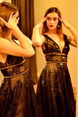 Beautiful young spectacular girl stands near the mirror in a shiny black dress with an open neckline 