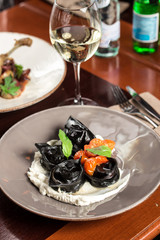 Traditional Italian black ravioli with cherry tomatoes and glass of white wine on wooden table at restaurant