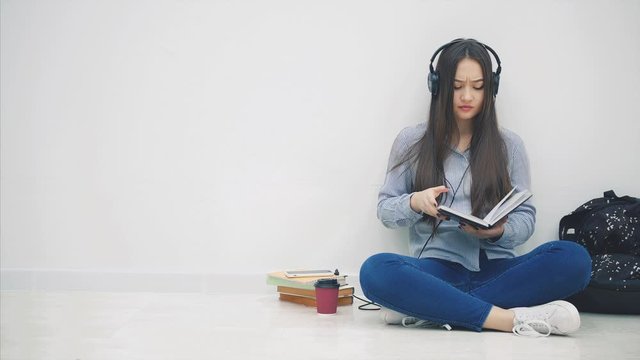 Funny young asian girl sitting on the floor in headphones, listening to the music, stops writing and takes selfie, posing, showing peace sign.