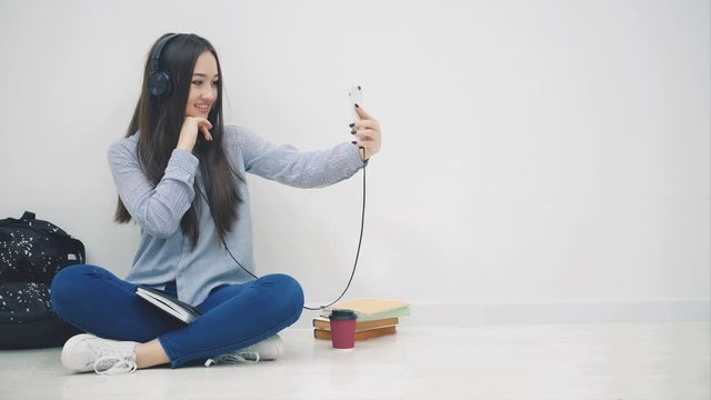 Funny young asian girl sitting on the floor in headphones, listening to the music, takes selfie, posing, waving hello, then messaging with someone.