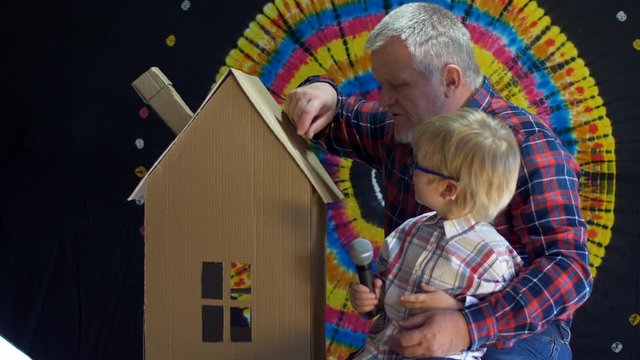 blond child and father with gray hair and beard in plaid shirts celebrate family holiday together. Father and son built cardboard house and examine it. family decides to place toys in it