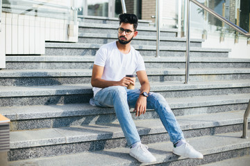 Young handsome indian man drink coffee while sitting on the stairs in the street