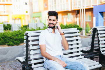 Young freelancer indian man sitting on bench, talking by phone, looking at camera. Handsome man in glasses wearing in white stylish shirt. It worker smiling, posing on background of modern city.