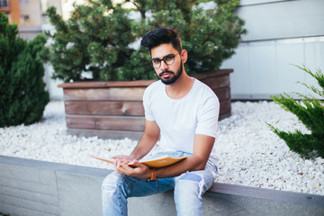 Obraz premium Young indian man in glasses read notebook sitting on the bench in the street