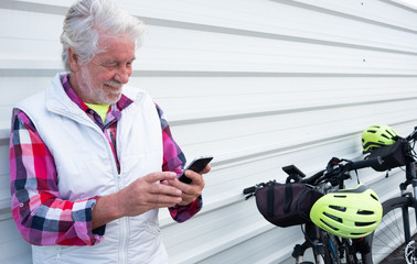 A smiling senior man with white hair and beard standing against a white metal wall. Looking at his mobile phone. Active people with his electric bicycle black color and yellow helmet