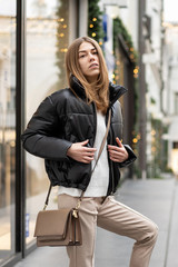portrait of a european young model in a warm leather jacket oversized with beige textile pants with a handbag on street in cloudy weather. Life style
