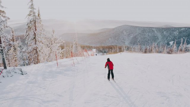 Skier in a red jacket on a snowy ski slope among the forest at sunset