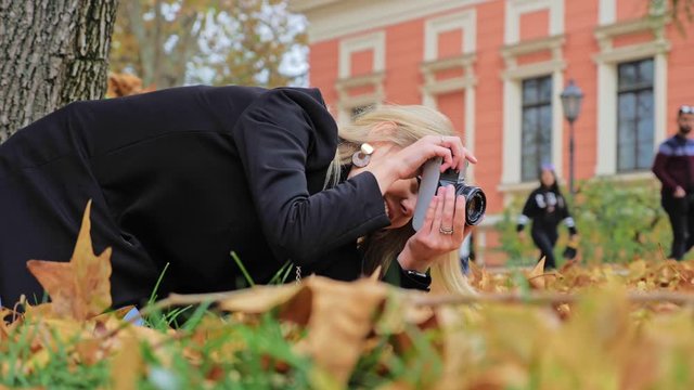 A young Caucasian female photographer takes autumn photos on a vintage retro film camera. Hipster takes pictures low near the ground, Controls his hand, asks passers-by to move away.