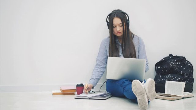 Cute asian girl sitting on the floor, involved in the process of preparation for exam, then raising up a sheet of paper with word EXAM written on it. Surrounded with learning supplies.