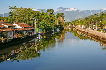 Fototapeta na wymiar View of river Perequê-Açu with houses, vegetation and rainforest mountains in background on a sunny day, reflections on the water in Unesco World Heritage town Paraty, Brazil