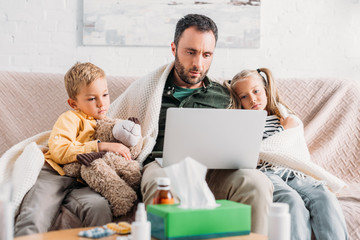 diseased children sitting on sofa near sick father using laptop