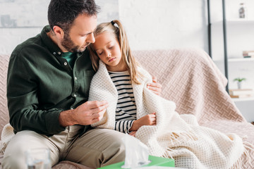 attentive father holding thermometer and hugging diseased daughter